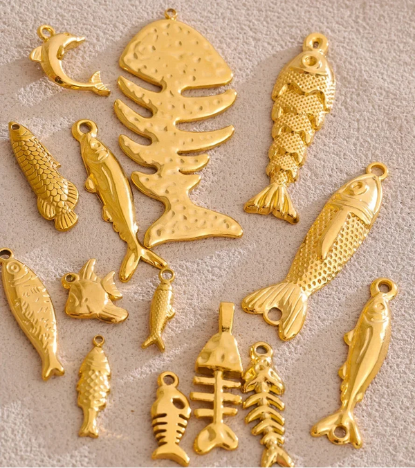 A set of gold-colored fishing-themed charms and pendants, including various fish, fish skeletons, and a small dolphin, arranged on a textured beige fabric background.