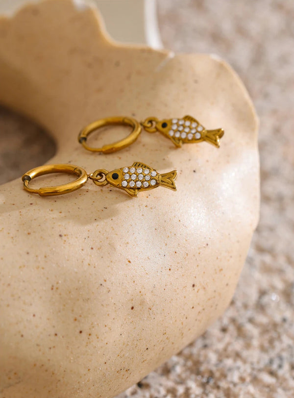 Gold fish-shaped earrings on a beige stone surface
