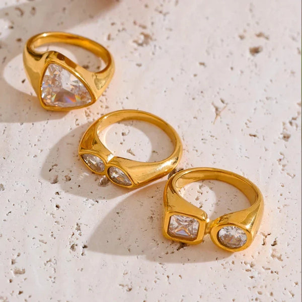 Three gold rings with clear gemstones on a textured beige surface