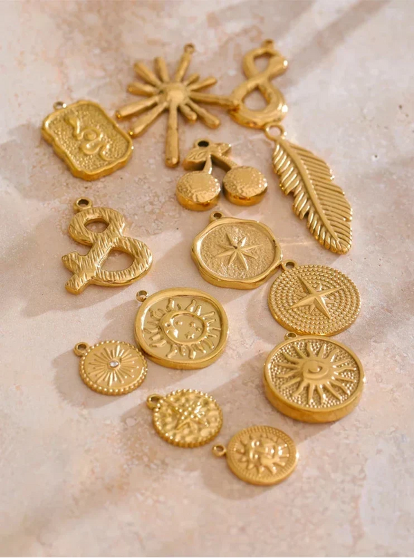 A collection of gold-toned metal jewelry charms and pendants with various abstract and symbolic designs, arranged on a light beige textured surface.