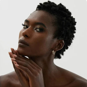 Woman wearing gold-plated hoop earrings, posing with hands folded under chin against neutral background.