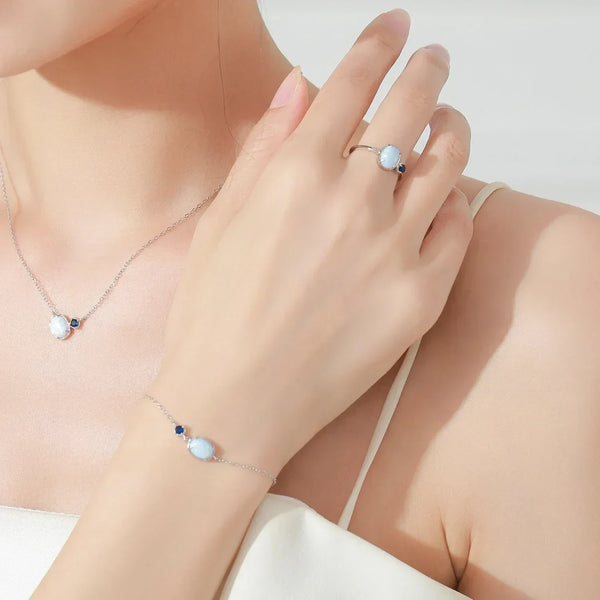 A close-up lifestyle shot of a model wearing a delicate silver jewelry set, including a necklace, bracelet, and ring, each featuring a light blue oval stone and a small dark blue accent.