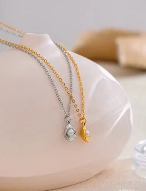 Two delicate shell-shaped pendant necklaces draped over a glossy white ceramic display bust. One necklace has a gold chain and a gold shell pendant with a white pearl, while the other has a silver chain and a silver shell pendant with a white pearl.