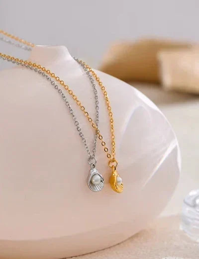 Two delicate shell-shaped pendant necklaces draped over a glossy white ceramic display bust. One necklace has a gold chain and a gold shell pendant with a white pearl, while the other has a silver chain and a silver shell pendant with a white pearl.