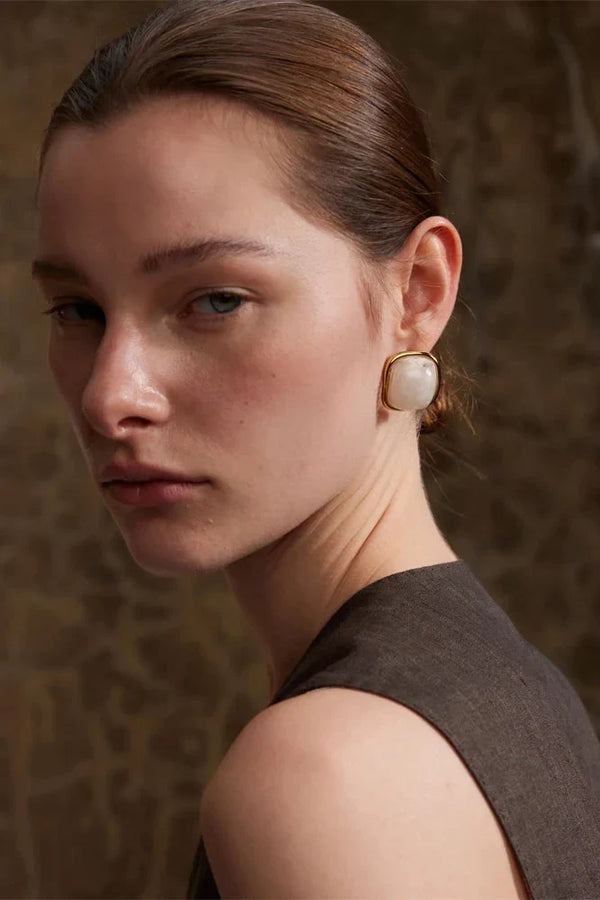Woman wearing a gold earring with a large pearl-like stone against a textured brown background