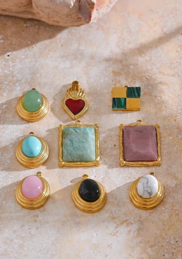A set of eight gold-tone fashion jewelry charms and pendants with various colorful gemstone and enamel inlays, arranged on a textured beige stone surface with the text 'FASHION ACCESSORIES' at the top.
