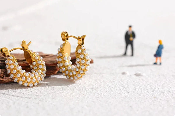 Gold-plated hoop earrings with white pearls on textured surface, miniature figures in background