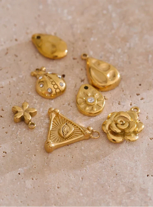 Six gold-toned jewelry charms with various organic and floral shapes, including a rose, triangle with sunburst, teardrop, and flower motifs, some adorned with small clear rhinestones, arranged on a textured beige stone surface.