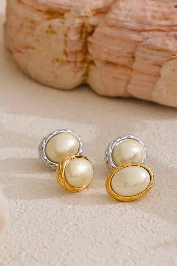 Pair of pearl stud earrings on a textured surface with seashells in the background.