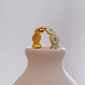 A gold ring featuring a series of white and yellow daisy flower charms, displayed on a light beige ceramic stand against a soft gray background.