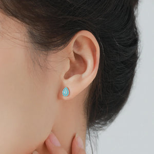 A close-up of a model's ear wearing a small teardrop-shaped stud earring with a light blue stone and a silver-toned border.