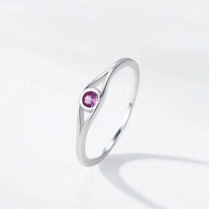 A silver ring with a unique curved triangular shank and a round pink gemstone in the center, resting on a light gray textured surface.