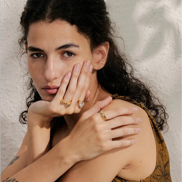 Woman with dark hair and tattoos on her arm, wearing gold rings, against a neutral background.