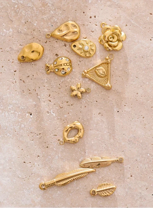 A set of nine gold-colored jewelry charms and pendants with various organic and geometric designs, displayed on a textured beige stone background.