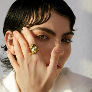 Woman wearing bold gold-plated statement ring, hand near face, white blouse visible