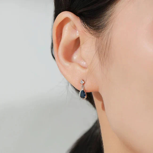 Teardrop-shaped earrings with a blue gemstone worn by a person against a neutral background