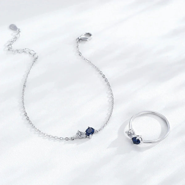 A silver bracelet and matching ring set on a light gray background. The delicate chain bracelet features a small oval blue gemstone and a tiny clear stone.