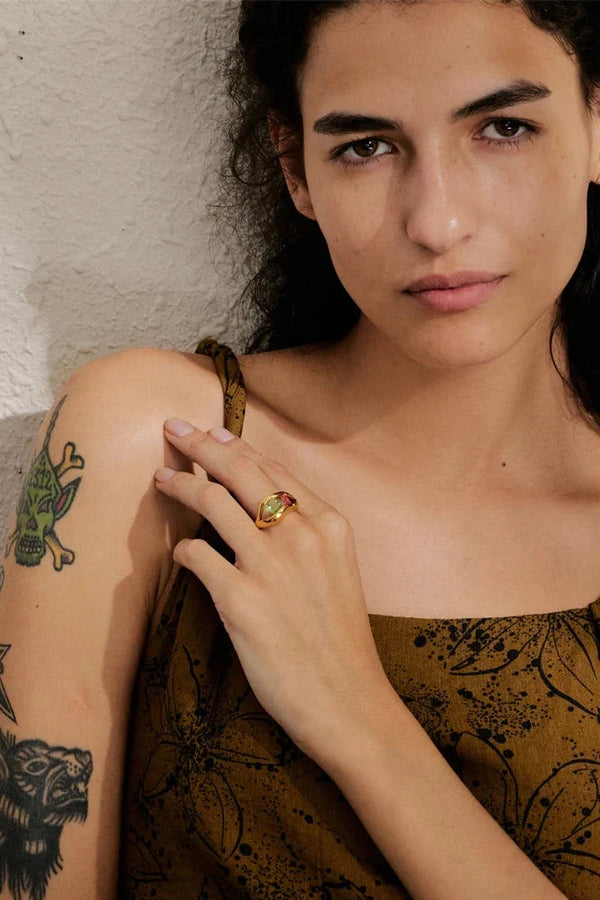 Woman with tattoos on her arm wearing a gold ring, against a neutral background