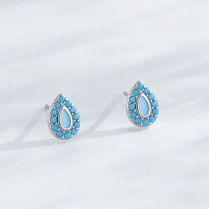A pair of silver teardrop stud earrings with a border of small turquoise-blue stones and a larger light blue teardrop center, displayed on a soft white textured surface.
