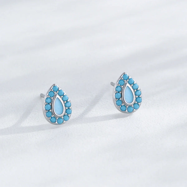 A pair of silver teardrop stud earrings with a border of small turquoise-blue stones and a larger light blue teardrop center, displayed on a soft white textured surface.
