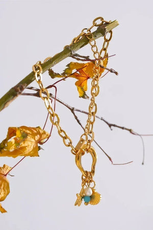 A gold-toned chain necklace with a shell-shaped charm, a small white pearl bead, and a turquoise accent, draped over a thin branch with dried yellow leaves against a light gray background.