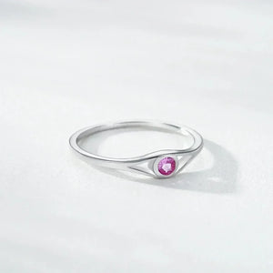 A silver ring with a unique curved triangular shank and a round pink gemstone in the center, resting on a light gray textured surface.
