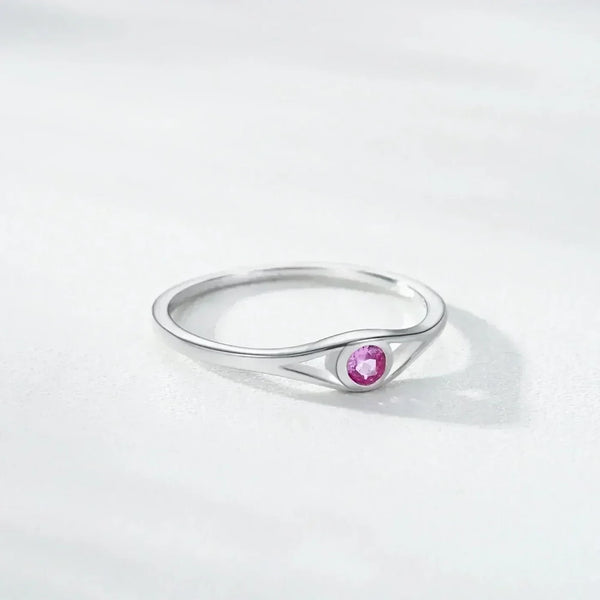 A silver ring with a unique curved triangular shank and a round pink gemstone in the center, resting on a light gray textured surface.