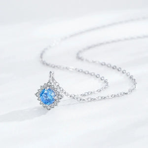 A silver chain necklace with a delicate snowflake-style pendant featuring a round-cut light blue gemstone surrounded by small clear stones, displayed on a soft white fabric background.
