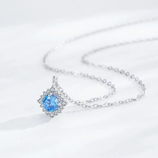 A silver chain necklace with a delicate snowflake-style pendant featuring a round-cut light blue gemstone surrounded by small clear stones, displayed on a soft white fabric background.