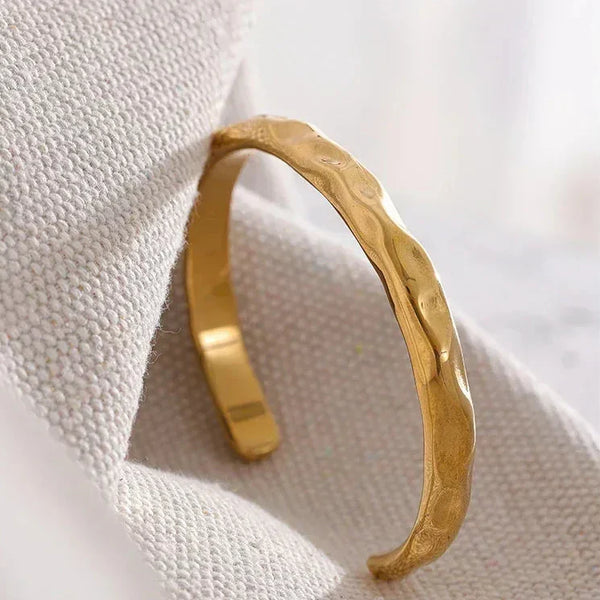 Textured gold-plated open bangle bracelet resting on a neutral fabric surface