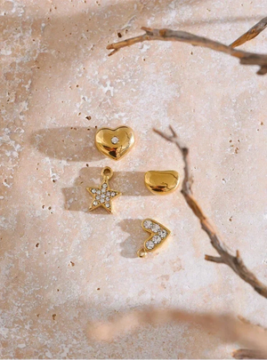 Gold jewelry pieces including a heart, star, and arrowhead on a textured beige surface with a branch.