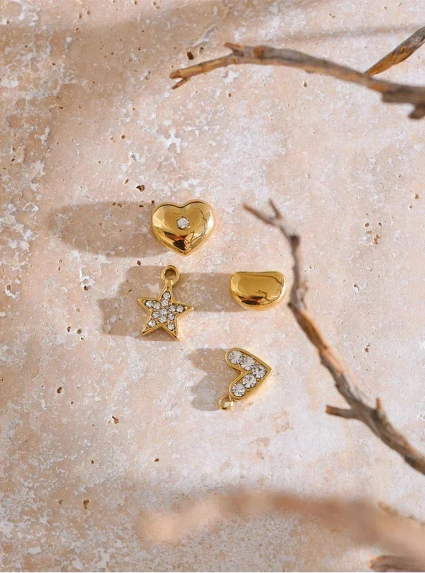 Gold jewelry pieces including a heart, star, and arrowhead on a textured beige surface with a branch.