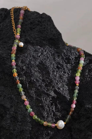 A multicolor beaded necklace with pink, green, orange, and brown round beads, a white pearl, and gold chain details, displayed on a textured black rock.