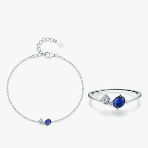 A silver bracelet and matching ring set on a light gray background. The delicate chain bracelet features a small oval blue gemstone and a tiny clear stone.