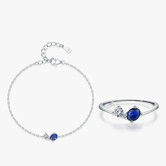 A silver bracelet and matching ring set on a light gray background. The delicate chain bracelet features a small oval blue gemstone and a tiny clear stone.