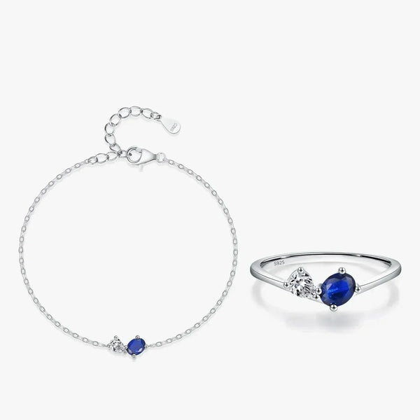A silver bracelet and matching ring set on a light gray background. The delicate chain bracelet features a small oval blue gemstone and a tiny clear stone.