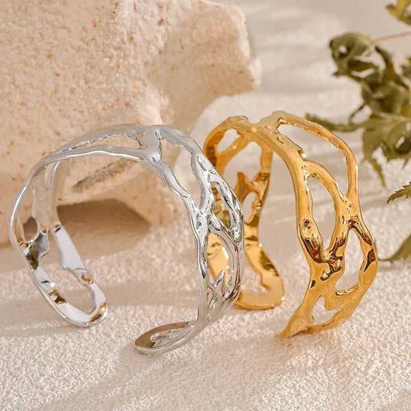 Two open cuff bracelets with an organic, branch-like organic pattern, one in polished silver and one in polished gold, displayed on a textured beige surface with a decorative shell and dried leaves.