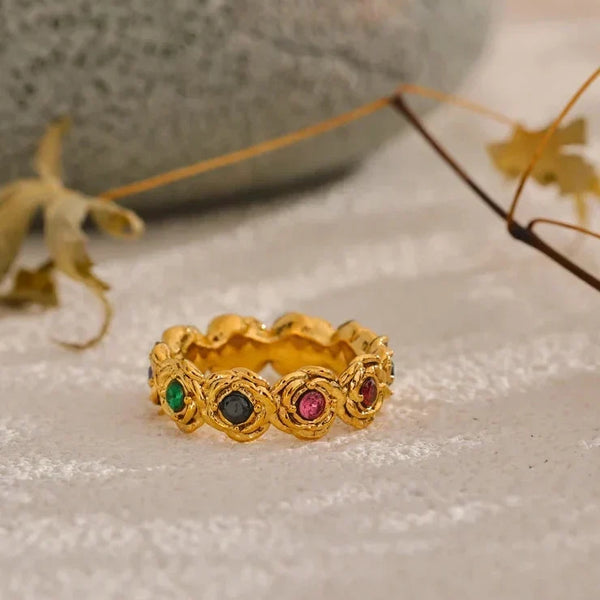 Gold ring with colored gemstones on a textured surface with leaves in the background