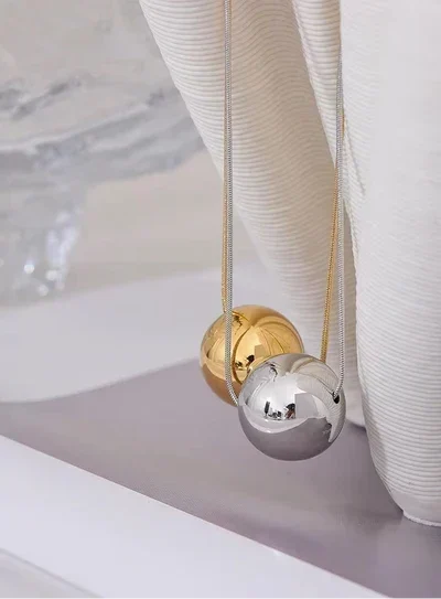 Two spherical pendant necklaces hanging from fine chains, one polished silver and one polished gold, displayed against a soft white ribbed fabric backdrop.