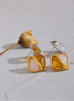 Gold and silver square-shaped earrings on a light gray surface with a dried flower.