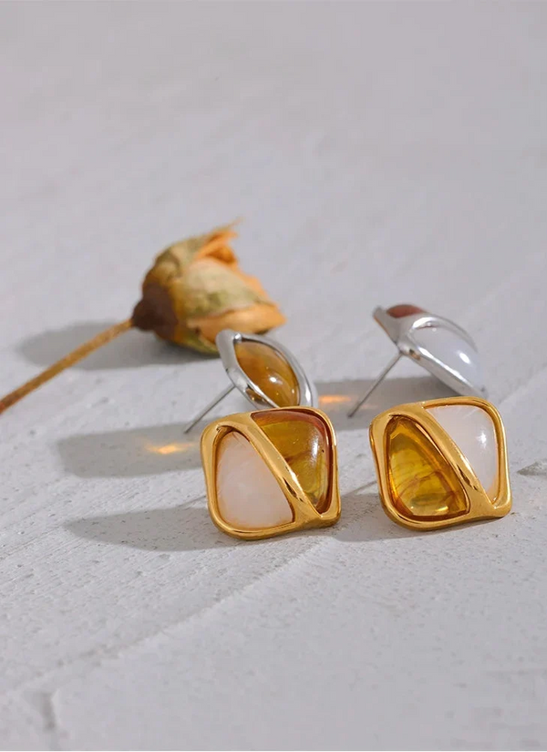 Gold and silver square-shaped earrings on a light gray surface with a dried flower.