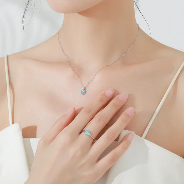 A close-up lifestyle shot of a model wearing a delicate silver necklace with a teardrop-shaped light blue pendant, paired with a matching silver ring on her finger.