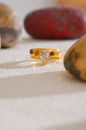 Gold ring with a clear gemstone on a textured surface with rocks in the background