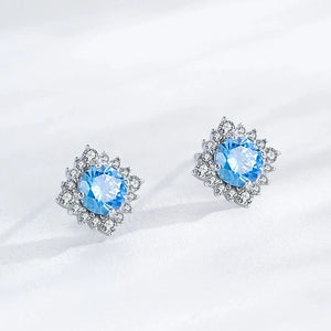 A pair of silver-toned stud earrings with a central light blue gemstone surrounded by small clear stones, displayed on a soft white fabric background.