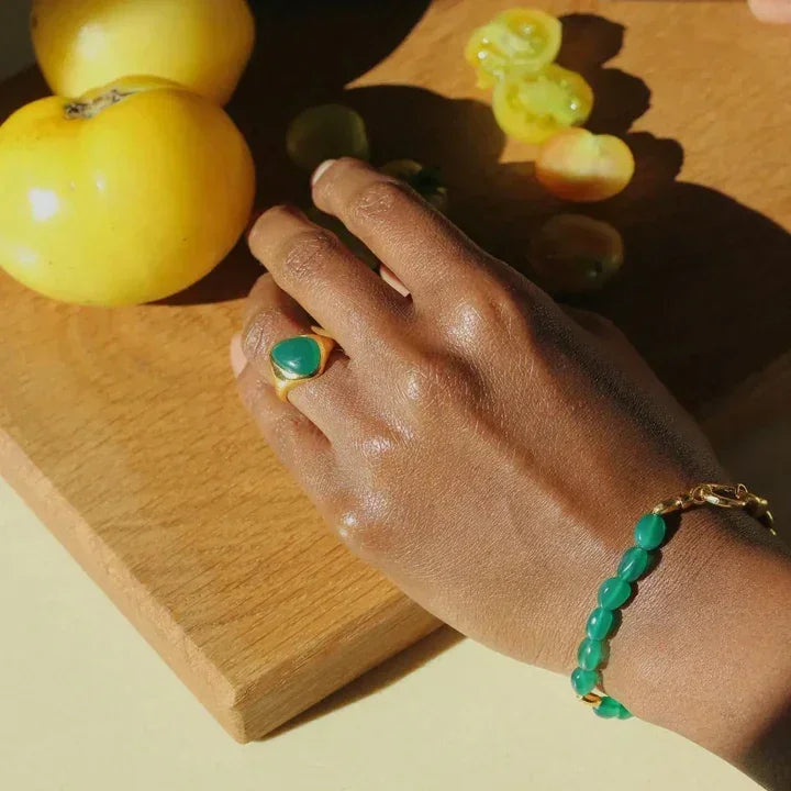 Hand wearing a green ring and bracelet with apples and grapes on a wooden board