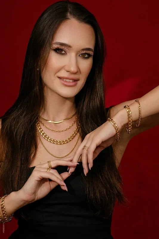 A model wearing a quality-crafted, layered gold chain necklace and bracelet set from the TM & Co. Jewels collection, set against a rich, editorial background.