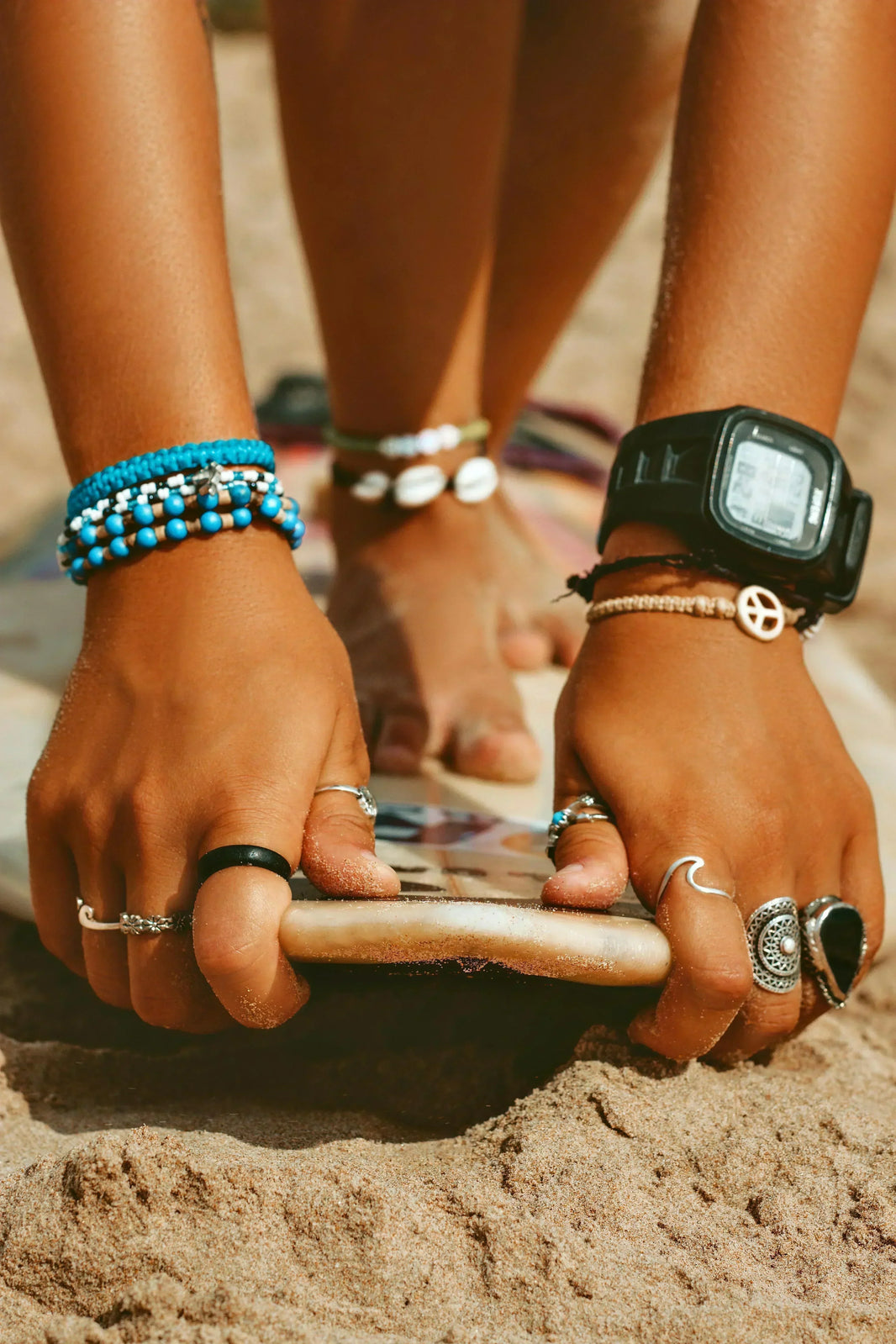Close-up of hands with multiple bracelets and rings on a sandy background
